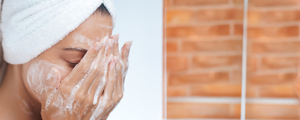 Mujer con una toalla en la cabeza lavando su rostro con un limpiador facial en su rutina de skincare