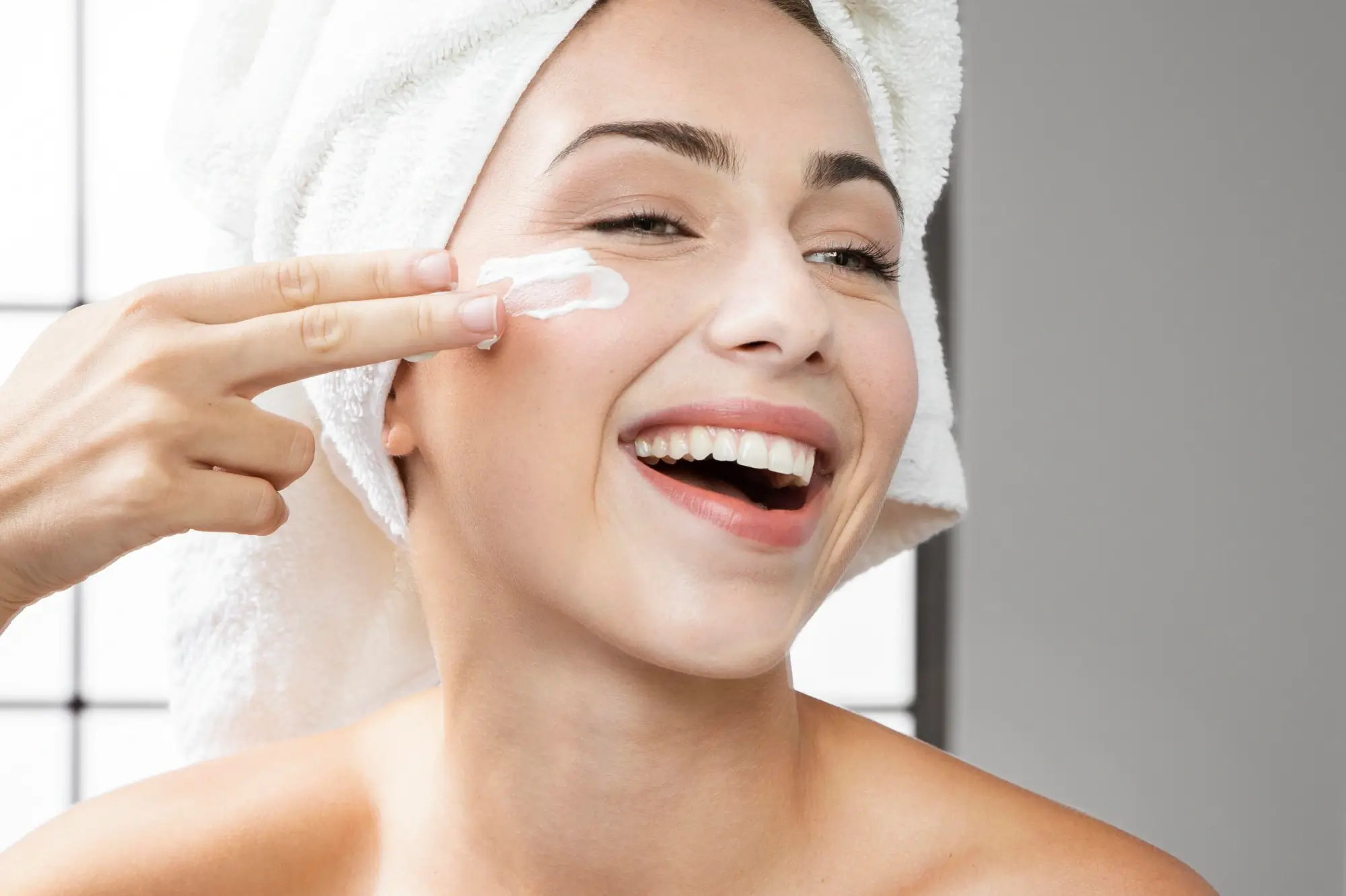 Mujer sonriente con toalla en la cabeza, aplicando crema en su rostro como parte de su rutina de hidratación facial
