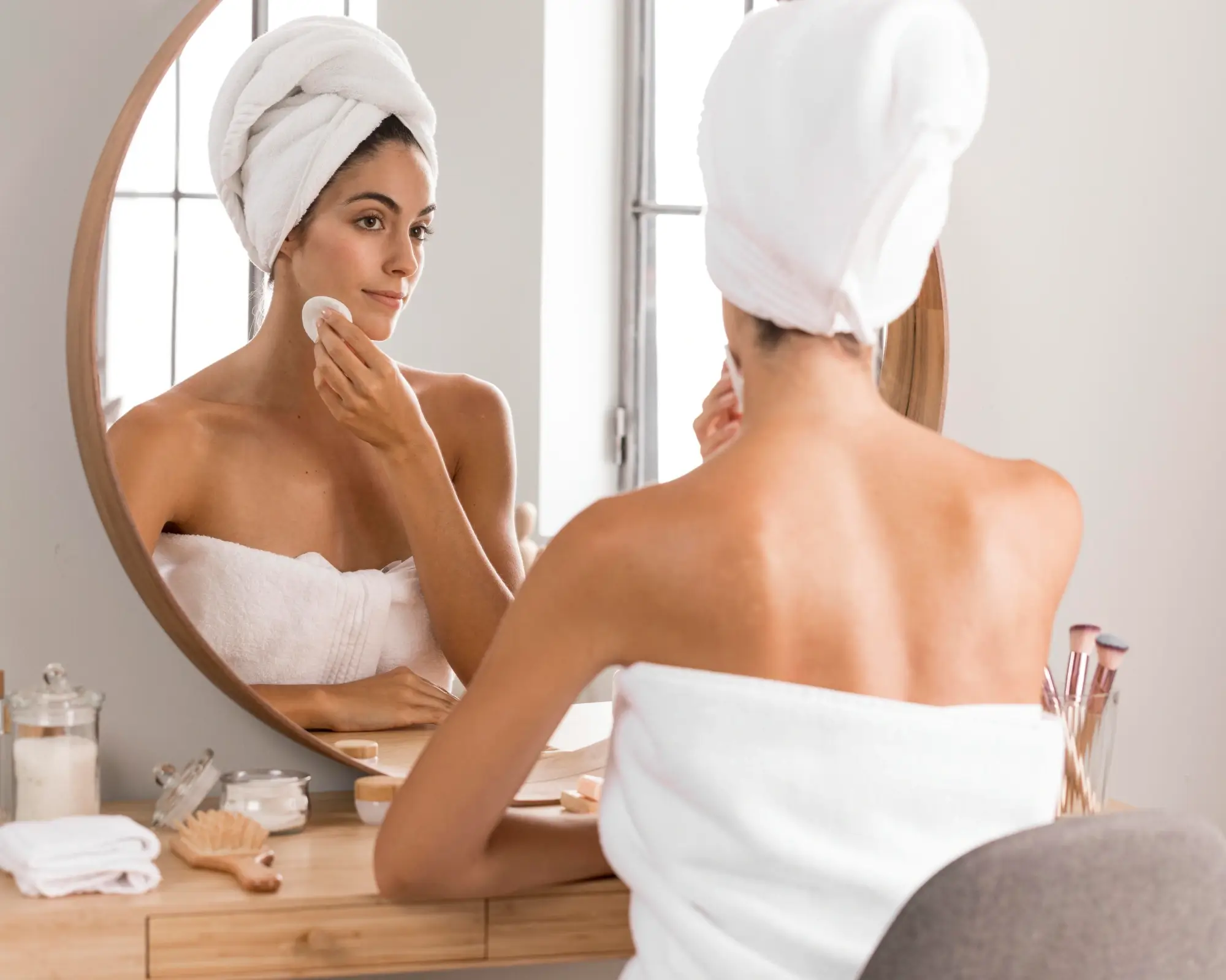 Mujer joven realizando su rutina de limpieza facial con un pad de algodón, frente a un espejo sobre un tocador de madera