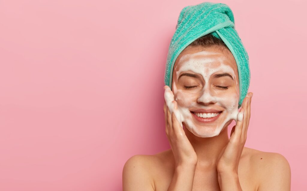 Mujer aplicando jabón facial en espuma sobre su rostro húmedo durante su rutina de limpieza facial.